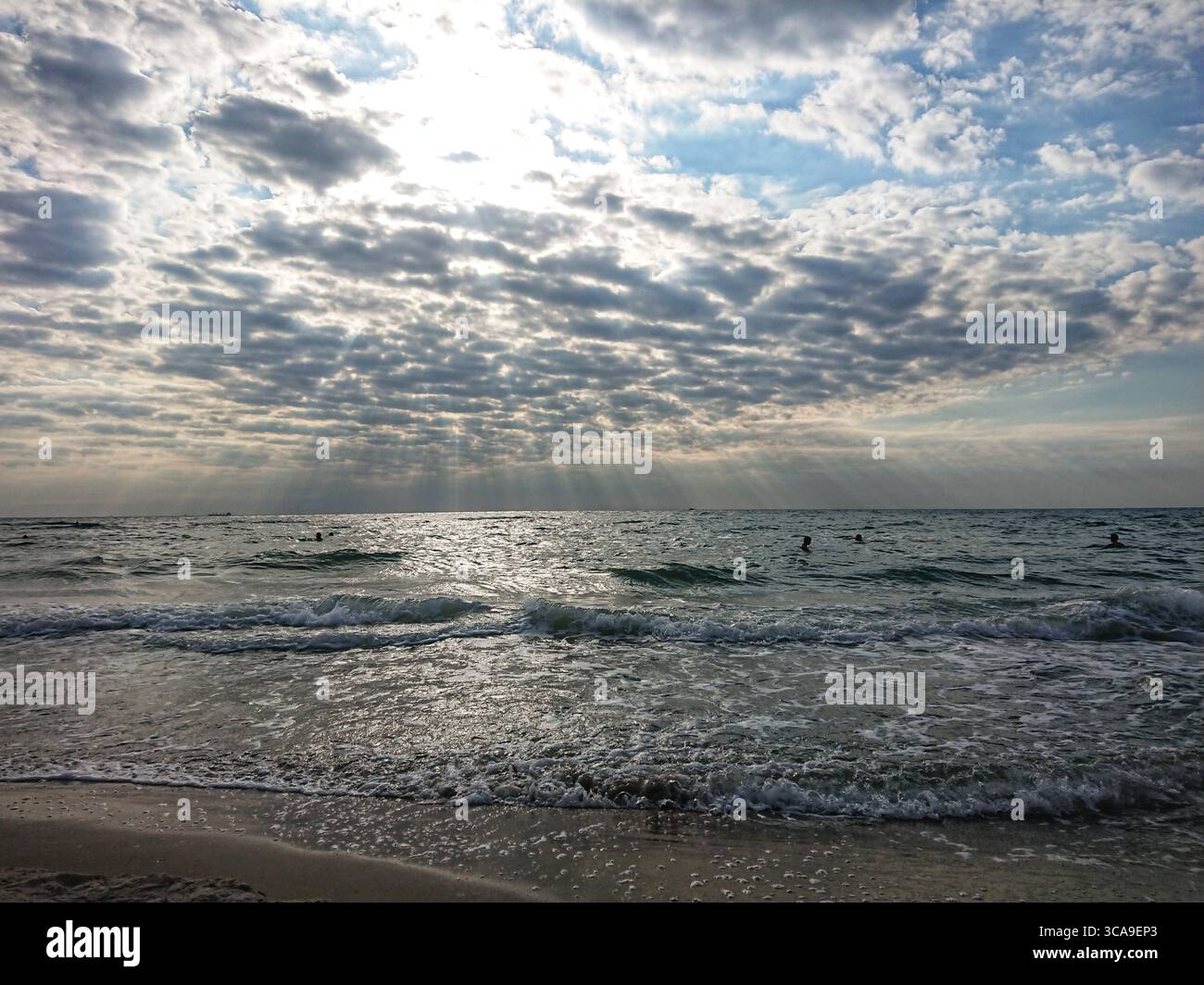 Wellen Rollen sanft auf einem Sandstrand unter dramatisch bewölktem Himmel während Sonnenuntergang am Meer, mit Schwimmen im Wasser, Sommer Küstenszene. Stockfoto