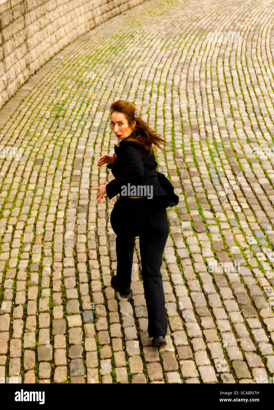 Eine Frau in einem schwarzen Anzug läuft einen Kopfsteinpflasterweg entlang und blickt über ihre Schulter zurück. Stockfoto