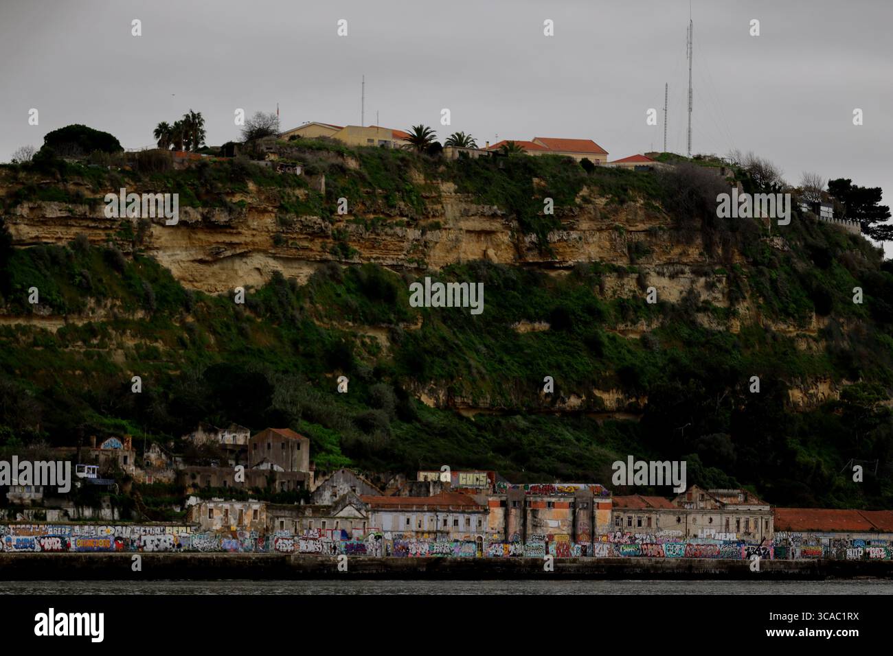 Grüne Klippe über graffiti markierten Häusern am Strand in Praia das Lavadeiras, Almada, Lissabon, mit einer mehrschichtigen urbanen Küste unter grauem Himmel. Stockfoto