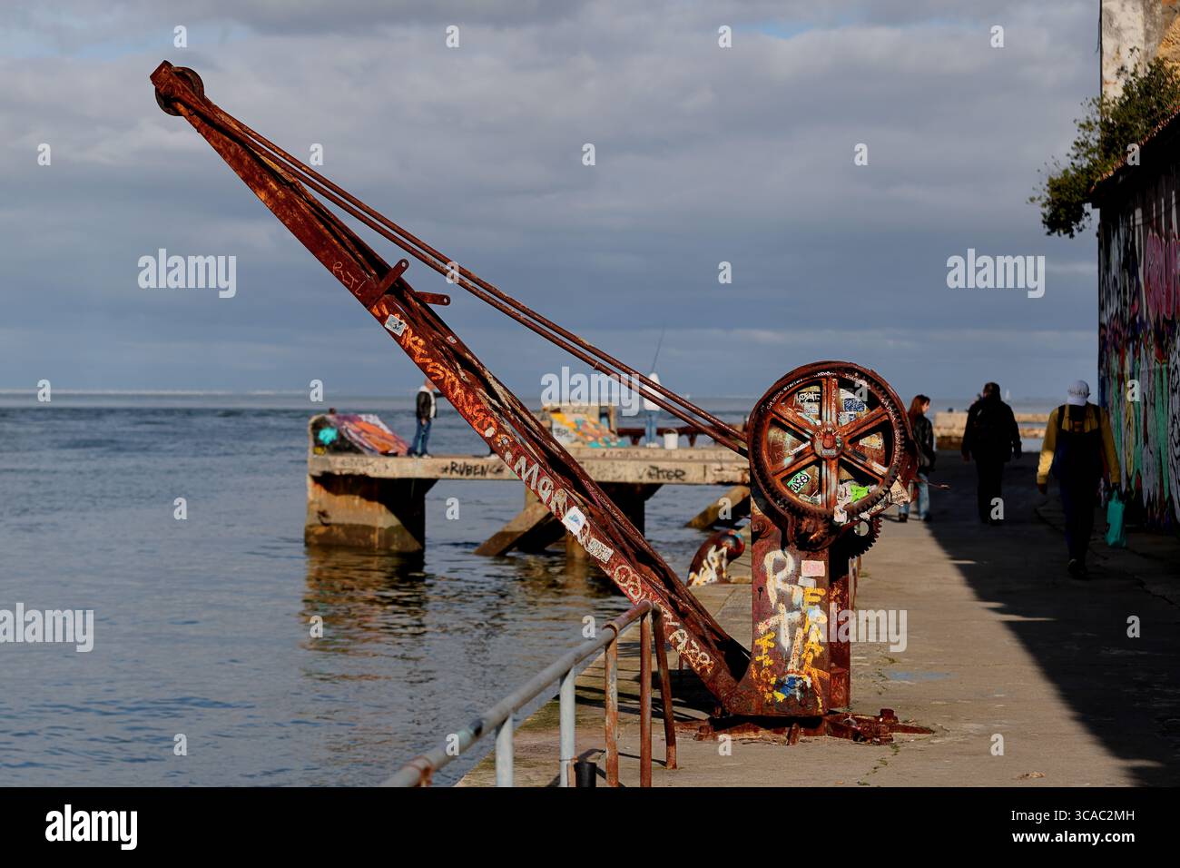 Rostiger Kaikran neben einer mit Graffiti gesäumten Mauer am Praia das Lavadeiras in Almada, Lissabon, blickt auf den Atlantik, während die Menschen den Pier unter Wolken schlendern. Stockfoto