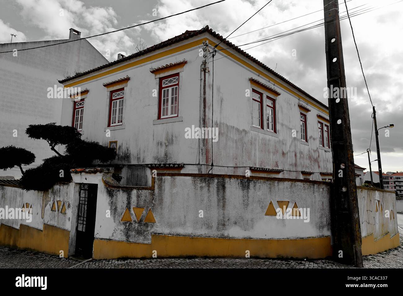 Eckhaus in Almada, Lissabon, mit rot getönten Fenstern, weiß getünchten Wänden und gelbem Sockel, eingerahmt von Versorgungsmasten und Drähten unter einem bewölkten Himmel. Stockfoto