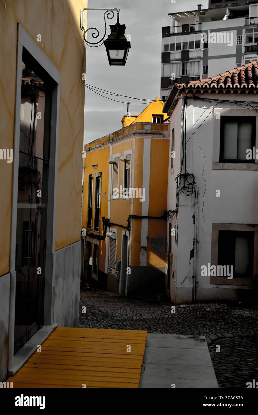 Enge Kopfsteinpflasterstraße in Almada, Lissabon, mit gelben und weißen Fassaden, die zu modernen Apartments unter einem dramatisch bewölkten portugiesischen Himmel führt. Stockfoto