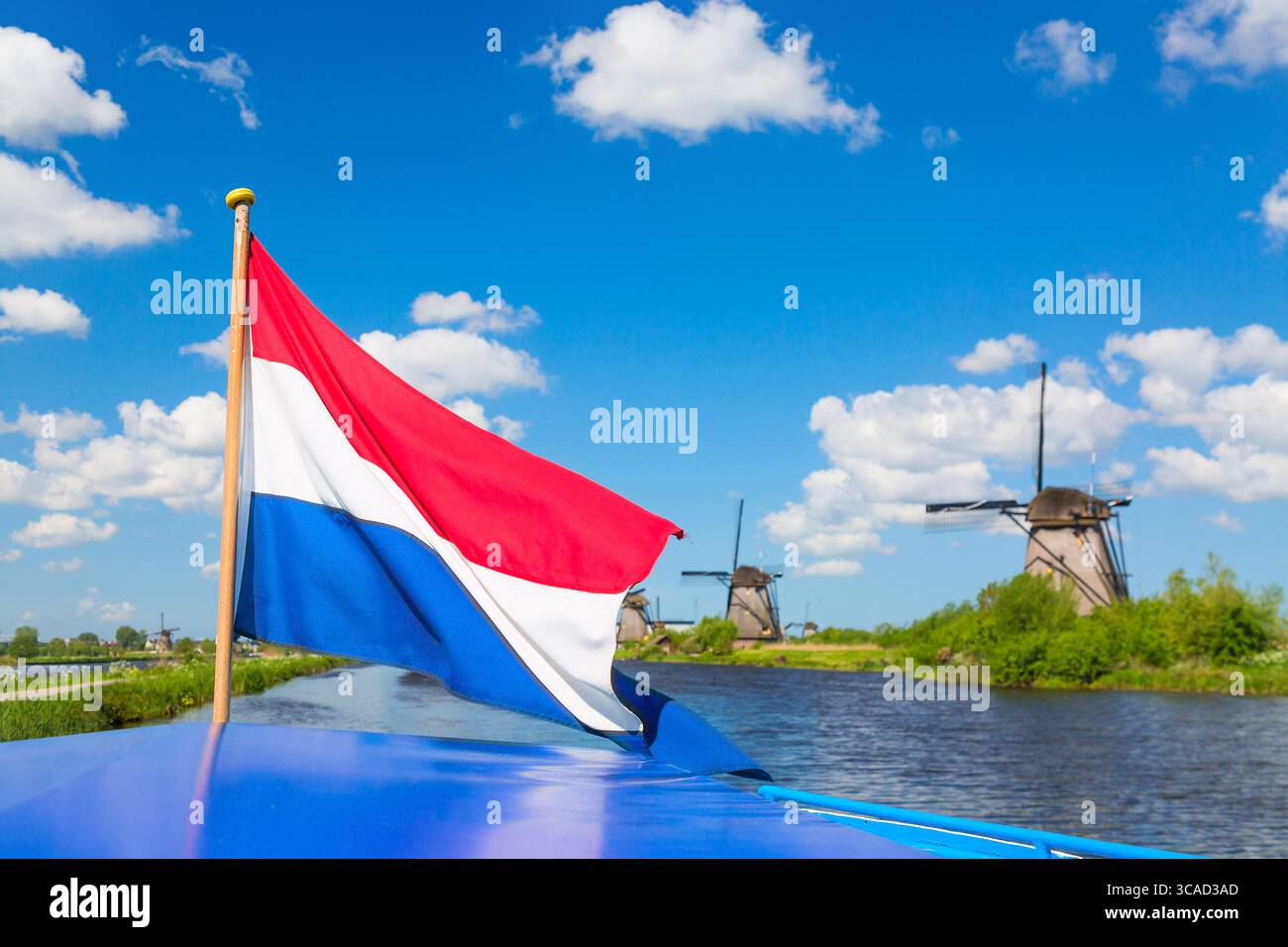 Die niederländische Flagge schwenkt auf einem Kreuzfahrtschiff gegen berühmte Windmühlen im Dorf Kinderdijk in Holland. Farbenfrohe ländliche Frühlingslandschaft in den Niederlanden, EU Stockfoto