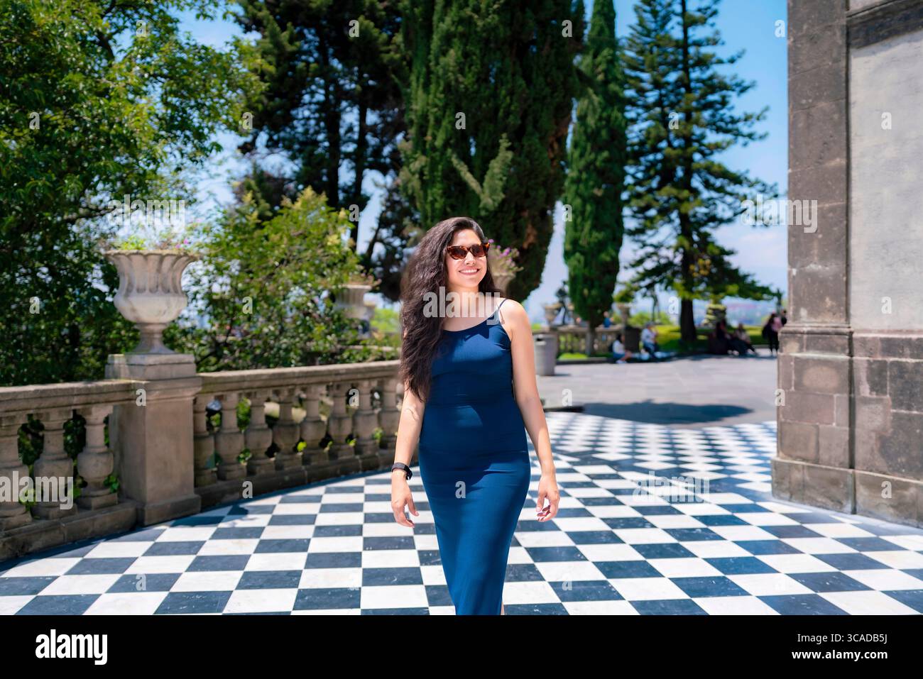 Fröhliche junge Latina mit langen welligen Haaren und Sonnenbrille geht mit einem Lächeln in einem blauen Kleid auf der ikonischen schwarz-weißen Terrasse von Chapultepec CA Stockfoto