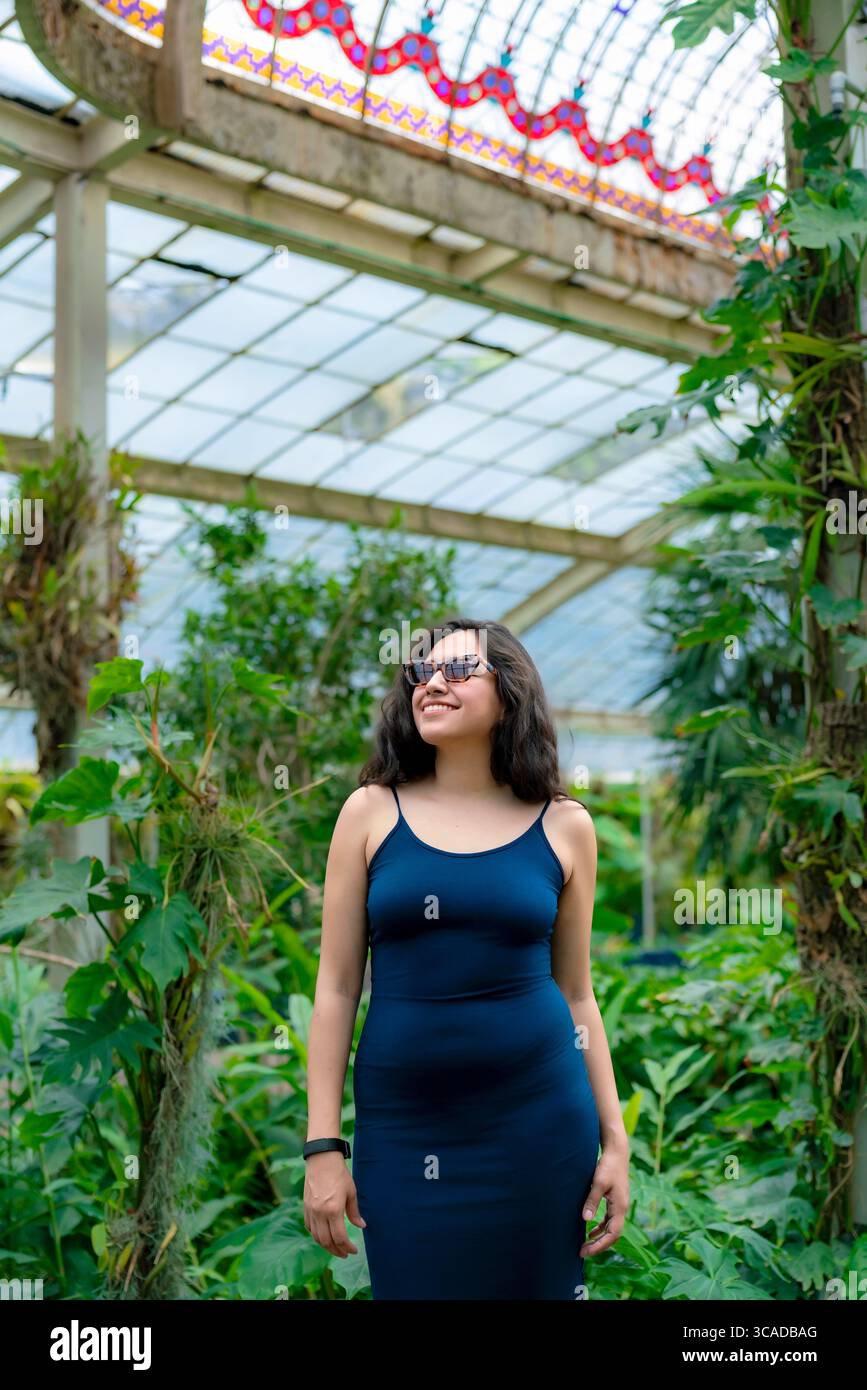 Die lächelnde junge Latina mit Sonnenbrille geht selbstbewusst durch den tropischen Innenbereich des Botanischen Gartens von Chapultepec, umgeben von exotischen Pflanzen Stockfoto