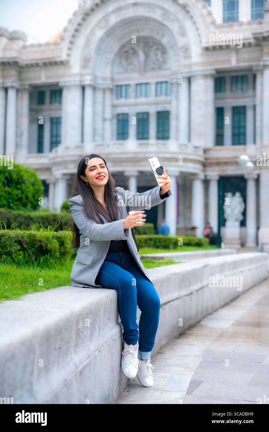 Selbstbewusste, glückliche junge Latina Geschäftsfrau, die lässig sitzt und lächelt, während sie ein Selfie in grauem Blazer und blauer Hose im Palacio de Bellas Artes macht. Stockfoto