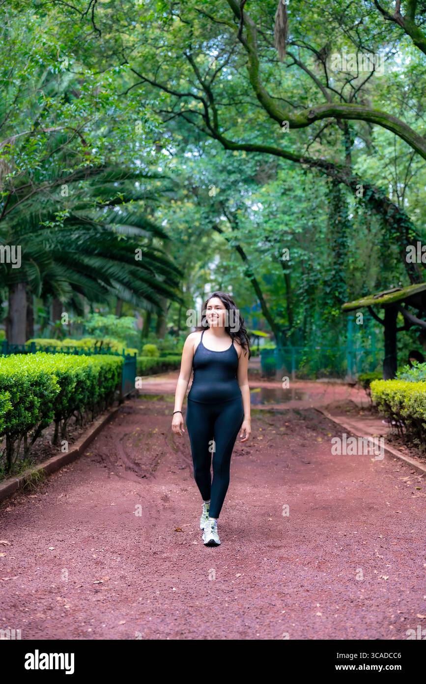 Sportliche junge Lateinfrau, die auf einem roten Kiesweg inmitten von Bäumen im Parque México läuft, einem berühmten grünen Platz in Condesa, ideal zum Joggen von Rela Stockfoto