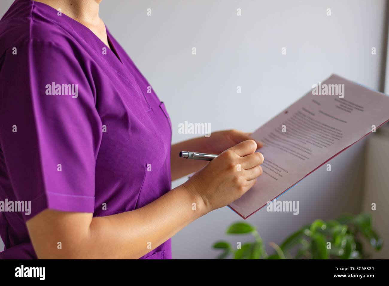 Weibliche Krankenschwester in lila Uniform liest und unterzeichnet ein medizinisches Dokument oder eine Akte, hält einen Stift in der Hand, steht in einer medizinischen Praxis Stockfoto