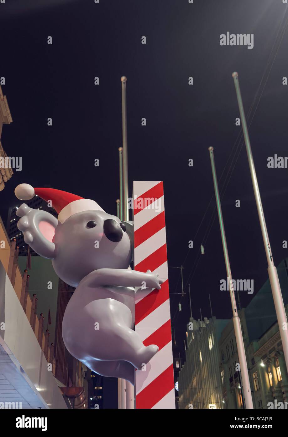 Riesiger aufblasbarer Koala mit weihnachtsmann-Hut, der nachts im Stadtzentrum von Melbourne einen Zuckerrohrstock klettert, mit Gebäuden im Hintergrund Stockfoto