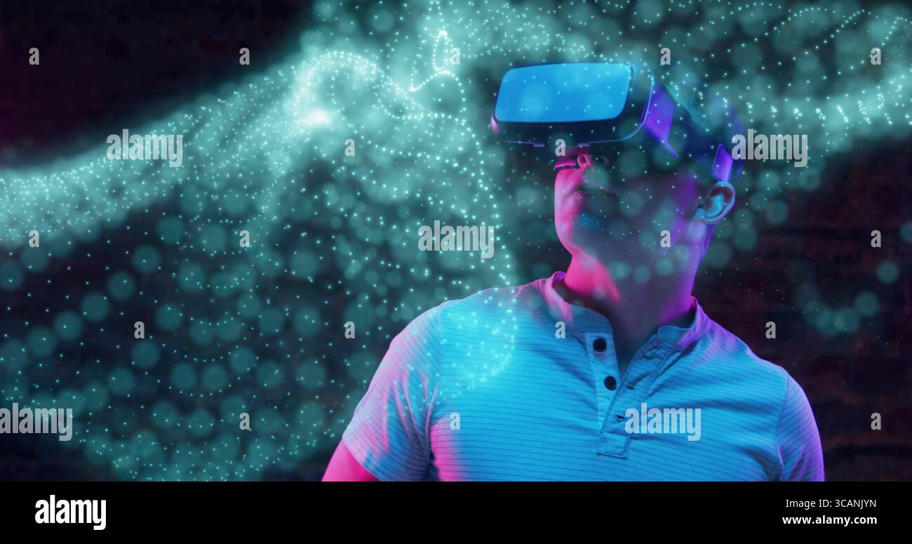 Stehender männlicher Erwachsener mit VR-Headset in dimmem Studio, mit leuchtenden digitalen Partikeln, Kopierraum Stockfoto