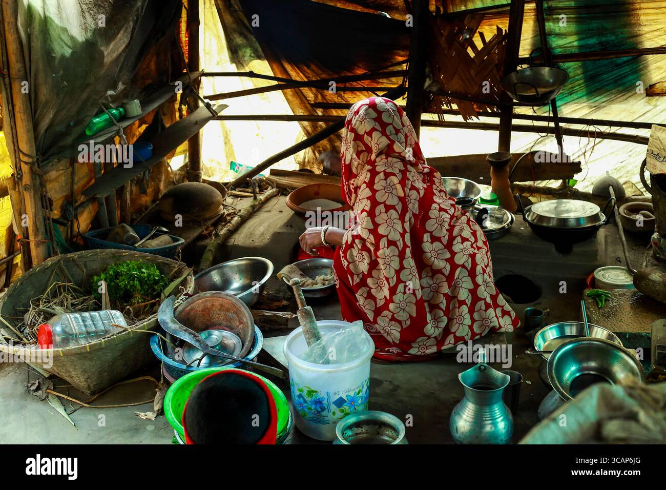 Cumilla, Bangladesch - 25. Januar 2020: Blick auf eine Frau in einem rot-weißen Sari, umgeben von rustikalen Küchenutensilien und bescheidenen Wohnungen, in weiches, diffuses Licht getaucht. Stockfoto