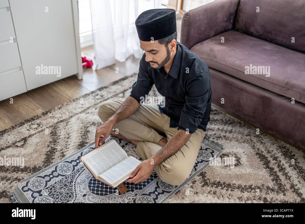 Muslimischer Mann liest den Koran, während er auf einer Gebetsmatte sitzt Stockfoto