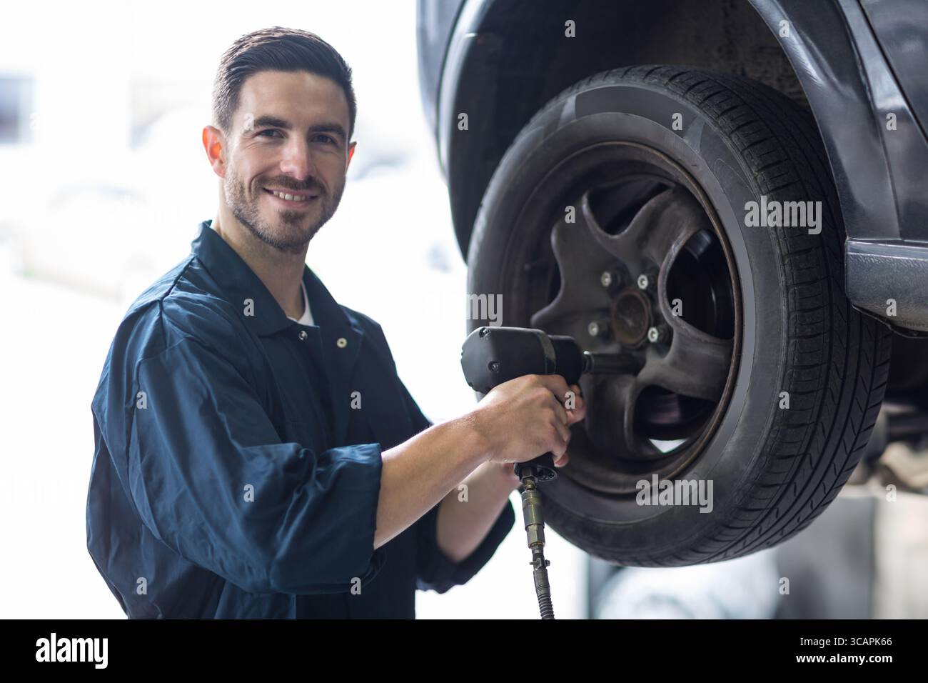 Männlicher Mechaniker, der neben dem angehobenen Autorad steht, entfernt die Radmuttern mit einem Druckluftschlüssel in der Garage Stockfoto