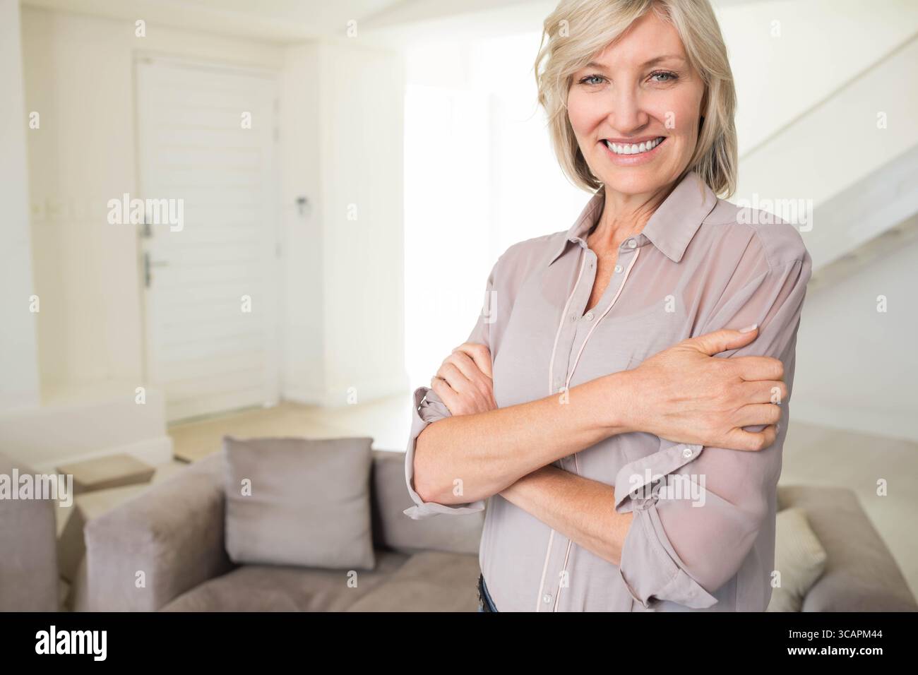 Reife Frau, die Arme überkreuzt, lächelnd in die Kamera im Wohnzimmer mit Sofakissen, Kopierraum Stockfoto