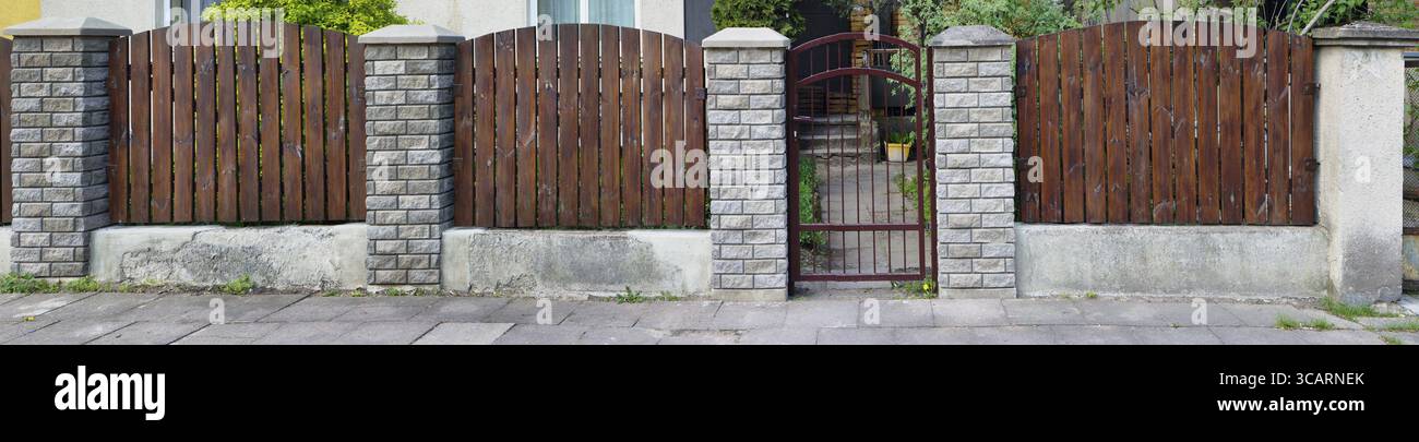 Der einfache Zaun des alten europäischen Hauses ohne Namen besteht aus vertikalen braunen, dünnen Kiefernplatten. Säulen aus einem dekorativen weißen Ziegel. Sonniger Frühlingstag Stockfoto
