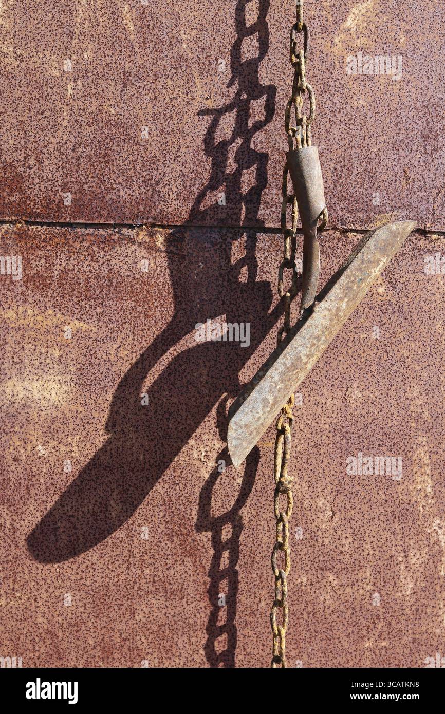 An der rostigen Metallwand des Dorfschuppen hängen an den Ketten alte alte landwirtschaftliche Werkzeuge. Sonniger Frühlingstag Stockfoto