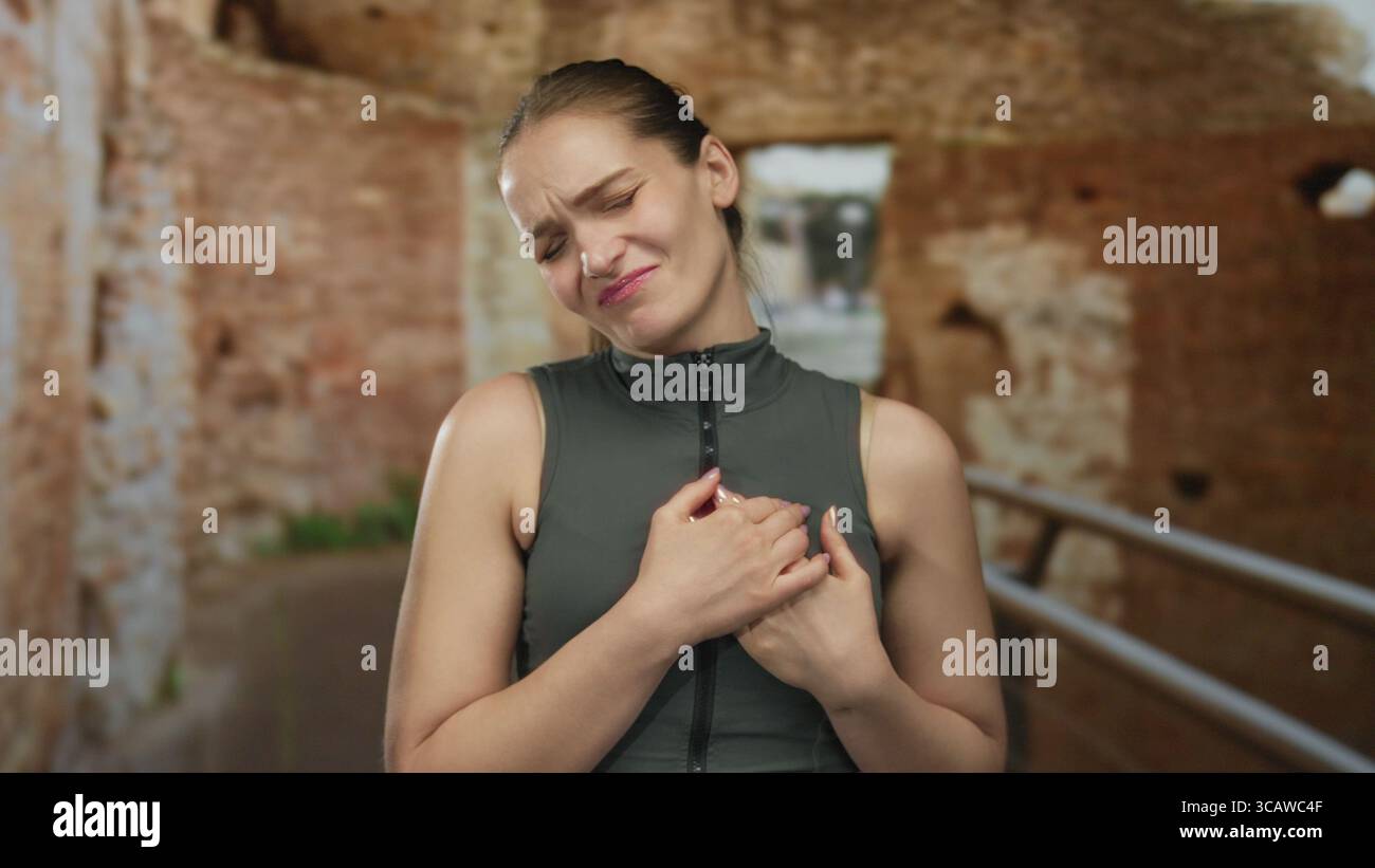 Eine grimmige junge kaukasische Frau, die die Brust an verwitterten antiken römischen Ruinen hält; Herzanfall-Schmerz. Stockfoto