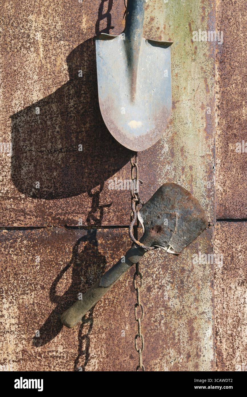 An der rostigen Metallwand des Dorfschuppen hängen an den Ketten alte alte landwirtschaftliche Werkzeuge. Sonniger Frühlingstag Stockfoto