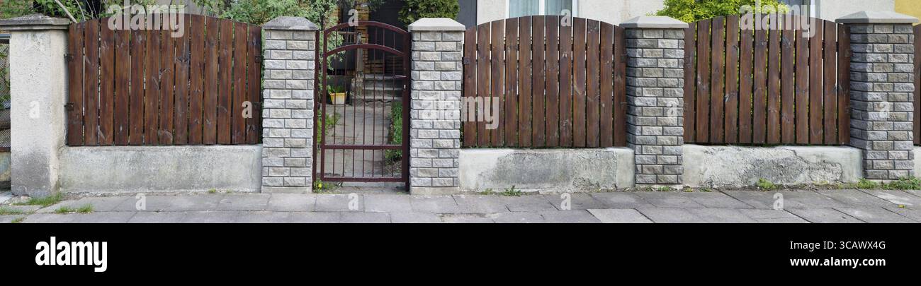 Der einfache Zaun des alten europäischen Hauses ohne Namen besteht aus vertikalen braunen, dünnen Kiefernplatten. Säulen aus einem dekorativen weißen Ziegel. Sonniger Frühlingstag Stockfoto
