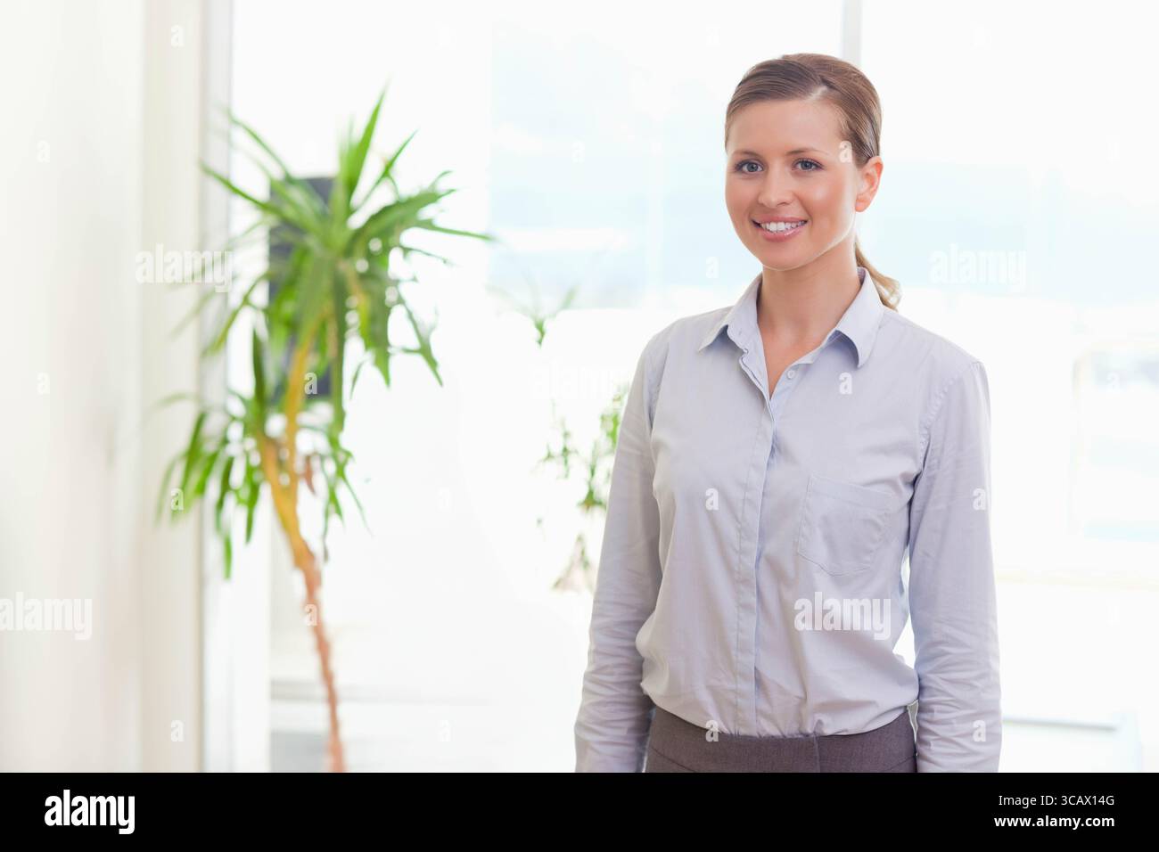 Frau, die bei einer Topfpflanze in der Nähe eines großen Fensters stand, trug ein blaues Hemd in einem hellen Büro, Kopierraum Stockfoto