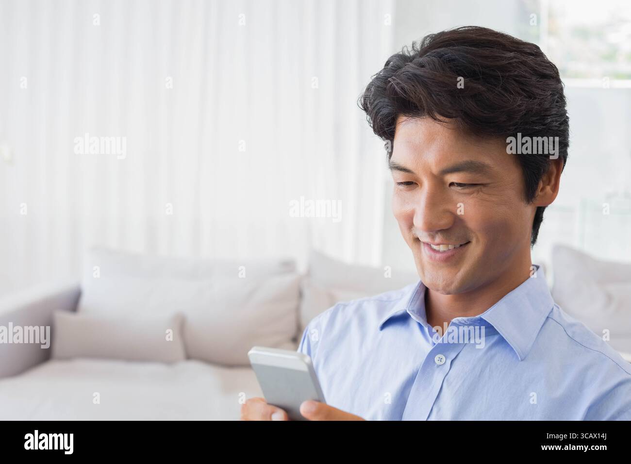 Japanischer Mann, der zu Hause das Smartphone auf dem Sofa hält, mit Kissen, Vorhängen, Kopierraum Stockfoto
