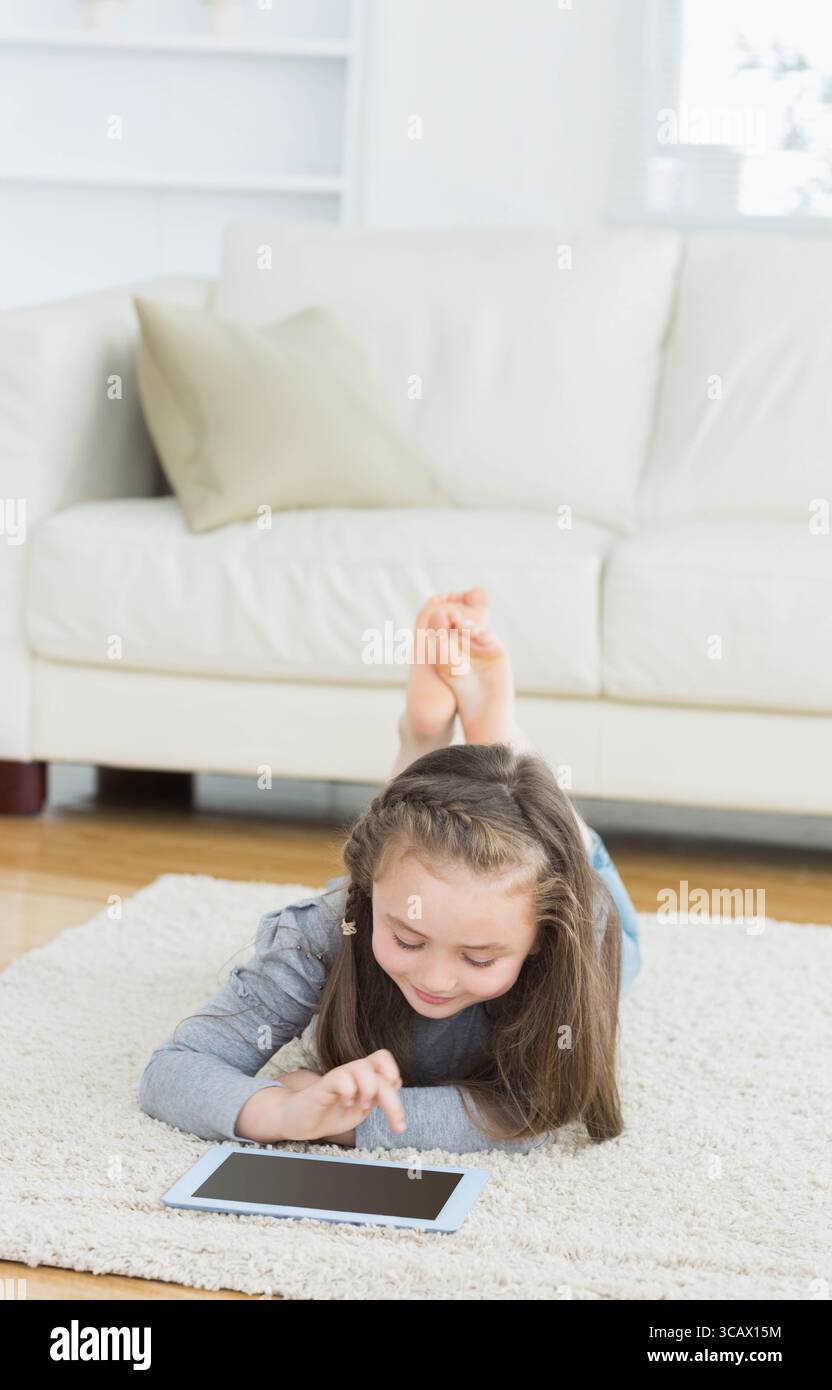 Mädchen, das zu Hause auf hellem Teppich liegend, das Tablet-Computer in der Nähe des weißen Sofas anklopft Stockfoto