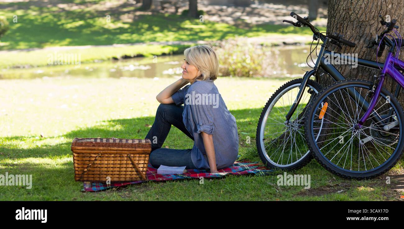 Erwachsene Frau sitzt auf karierter Decke am Teich im Park mit Picknickkorb und Fahrrädern Stockfoto