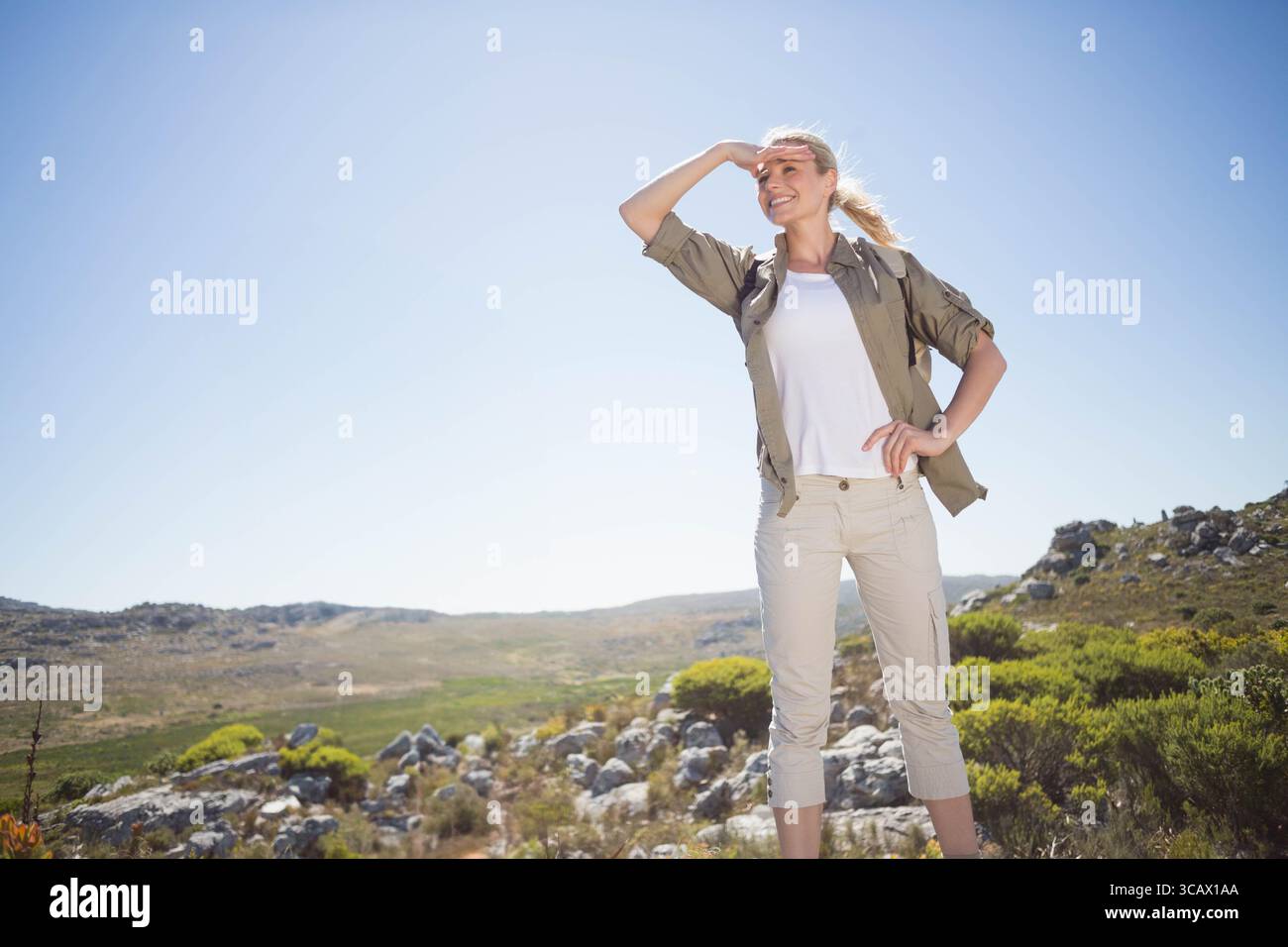 Der Wanderer steht auf felsigen Hügeln und schattet die Augen unter klarem Himmel und blickt mit seinem Rucksack über das Buschland Stockfoto