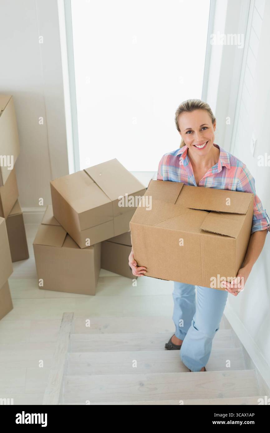MittelErwachsene Frau, die eine helle Holztreppe in einem hellen Eingangsbereich klettert und einen Karton trägt Stockfoto