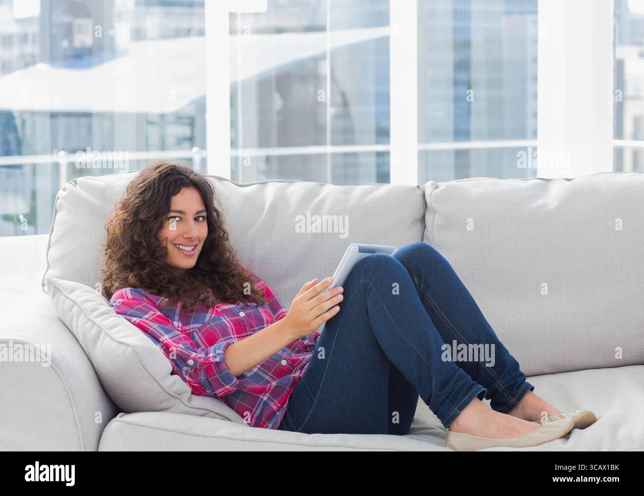Frau, die sich auf einem hellen Sofa im modernen Wohnzimmer mit einem Tablet zurücklehnt und die Skyline der Stadt zeigt Stockfoto