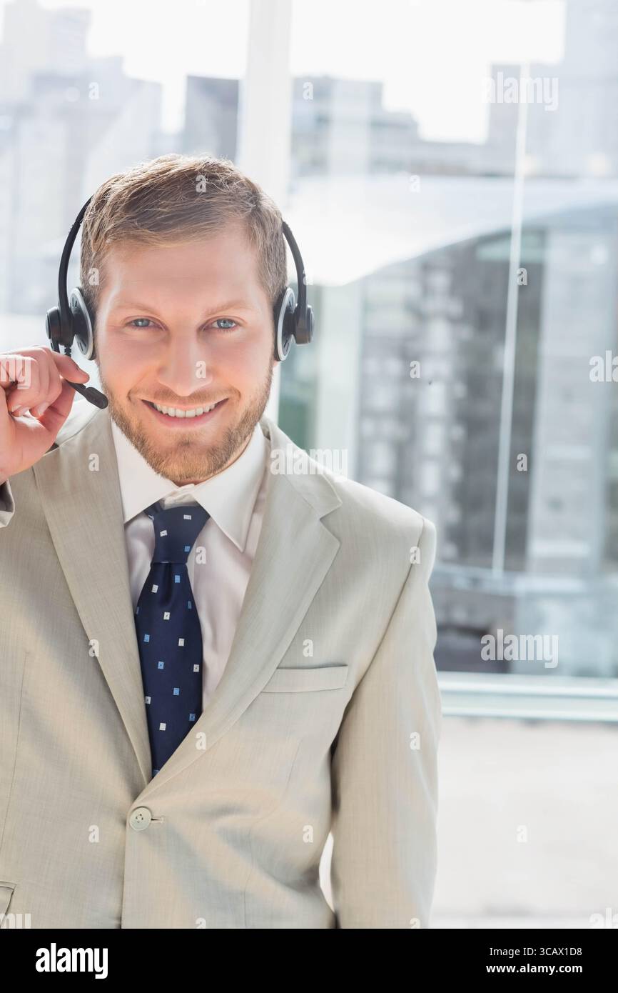 Mann mit Anzugsjacke und Headset-Mikrofon, stehend am Bürofenster mit Blick auf den Fluss und die Skyline Stockfoto