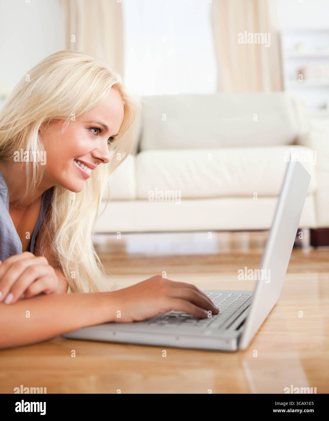Blonde Frau, die auf dem Boden liegt und auf silbernem Laptop neben dem hellen Sofa im Wohnzimmer tippt Stockfoto