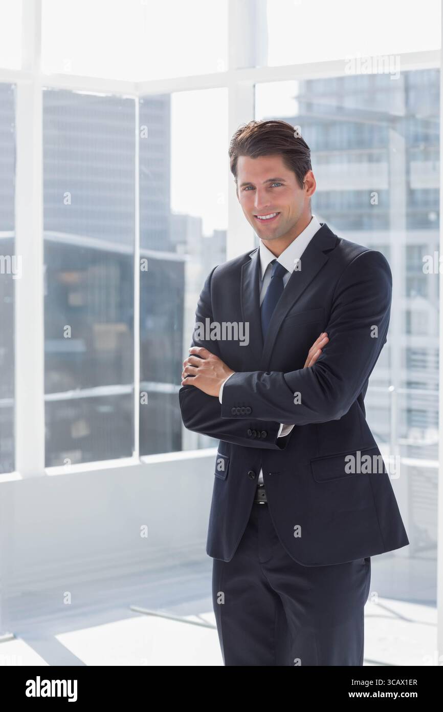 Männlicher Profi, der mit überkreuzten Armen im Business-Anzug im Hochhaus an Glasfenstern steht Stockfoto