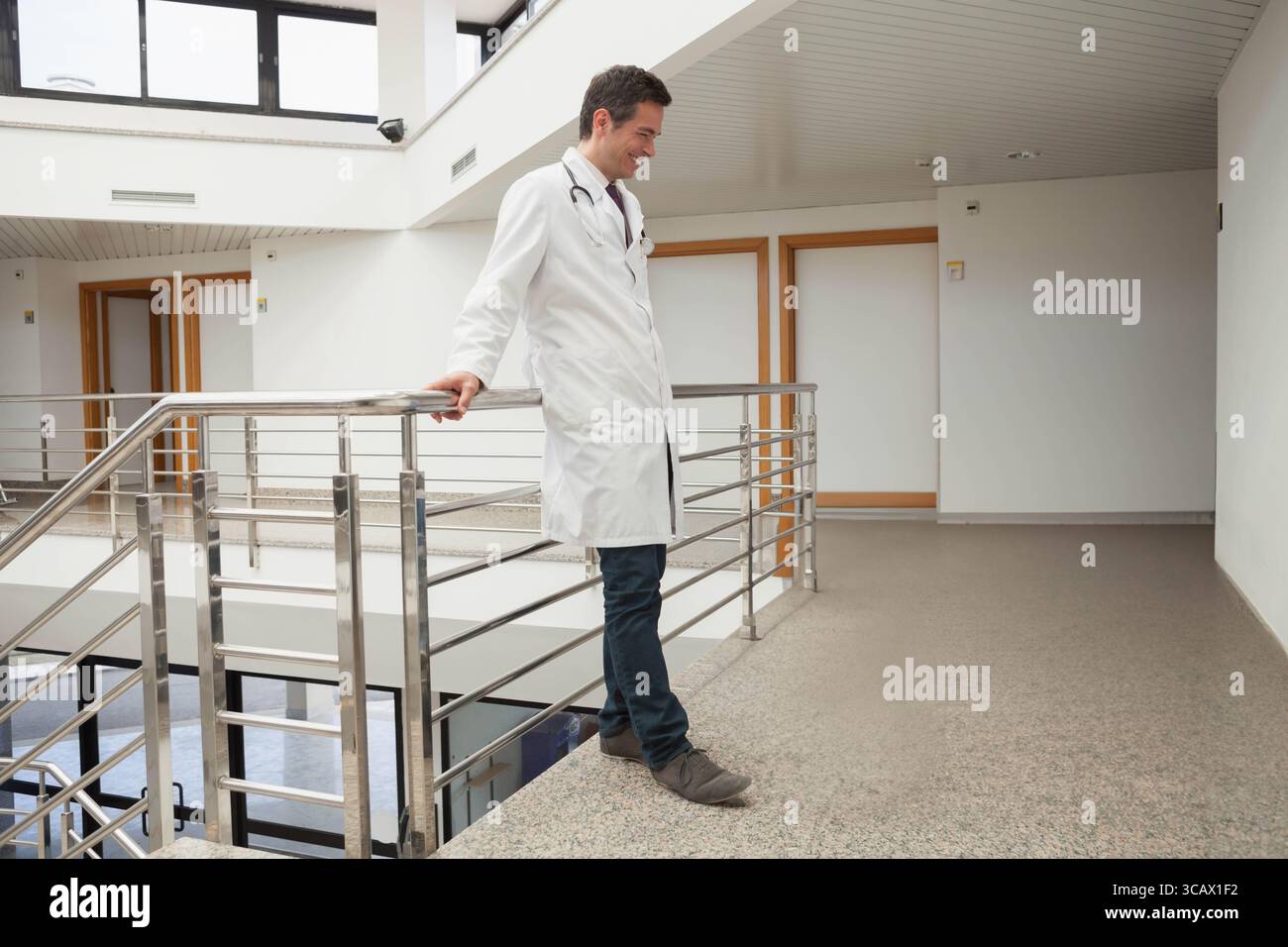 Männlicher Arzt mit Laborkittel und Stethoskop, gestützt auf Geländer im Krankenhausatrium, Kopierraum Stockfoto