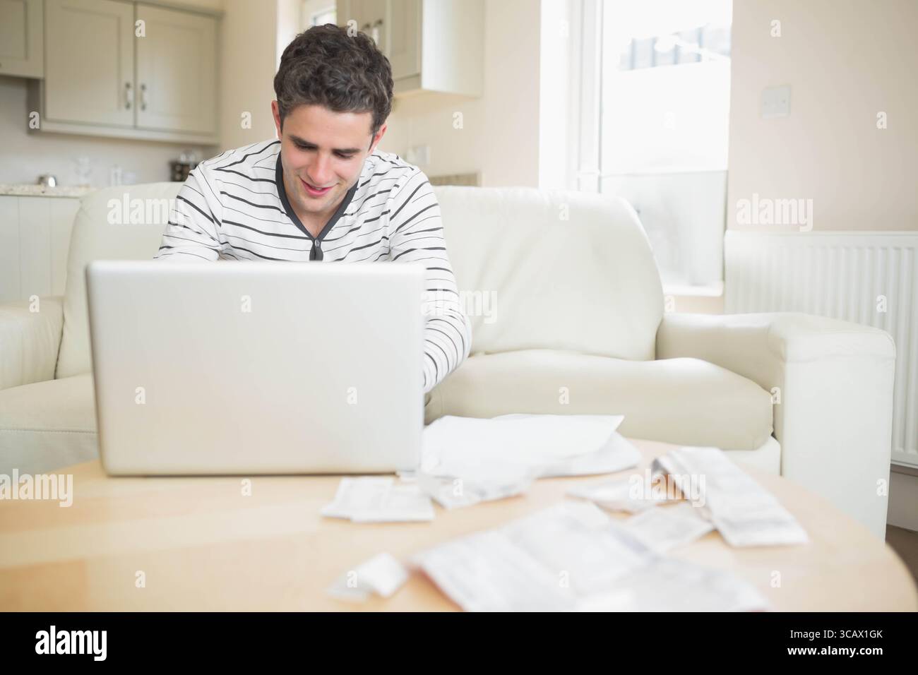 Ein hellhäutiger Mann sitzt zu Hause und arbeitet auf silbernem Laptop an einem hölzernen Kaffeetisch mit Papieren Stockfoto