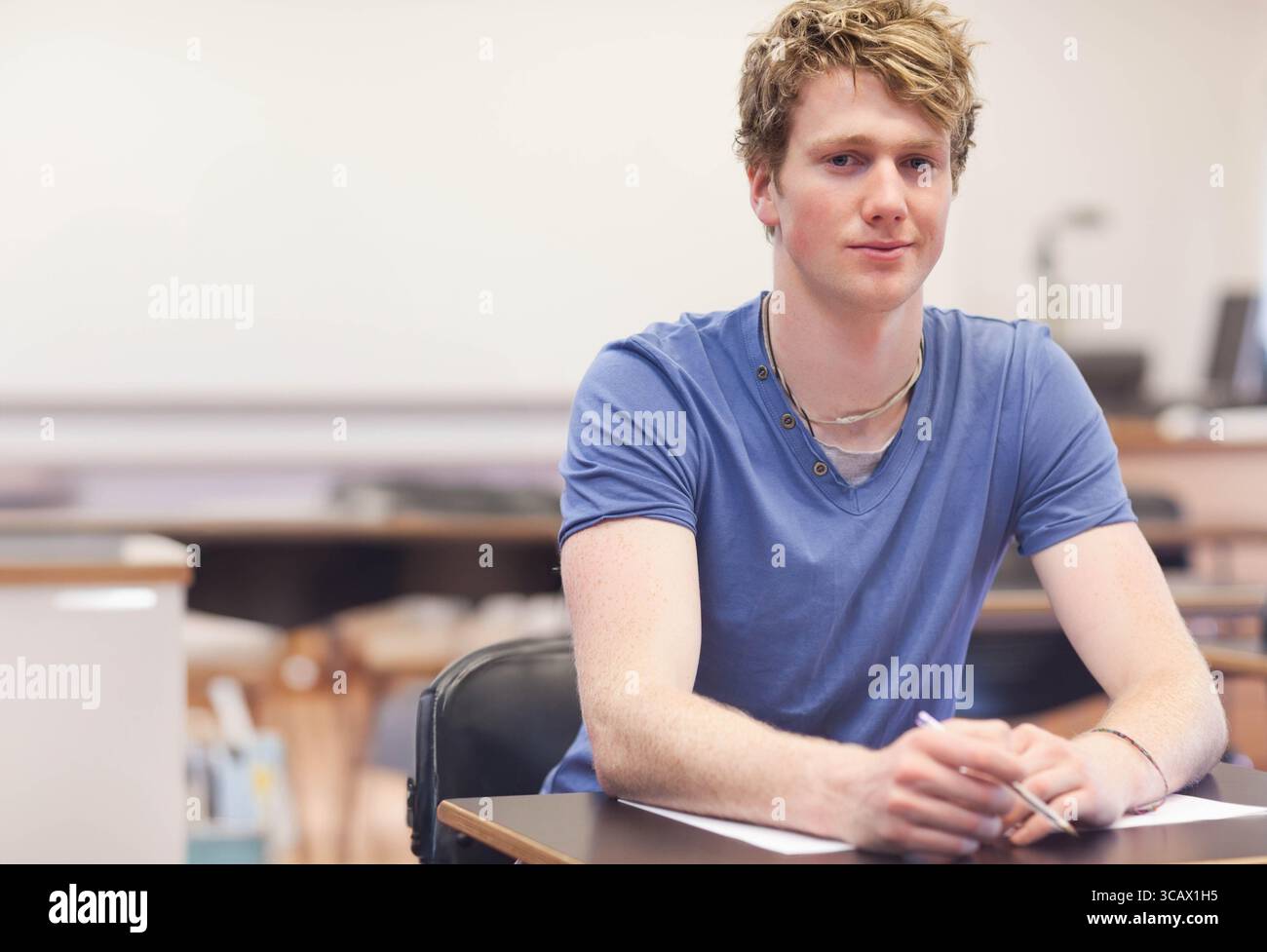 Männlicher Schüler mit blauem Hemd, der auf einem Blatt Papier schreibt, am Schreibtisch im Klassenzimmer mit Stift-Monitor Stockfoto