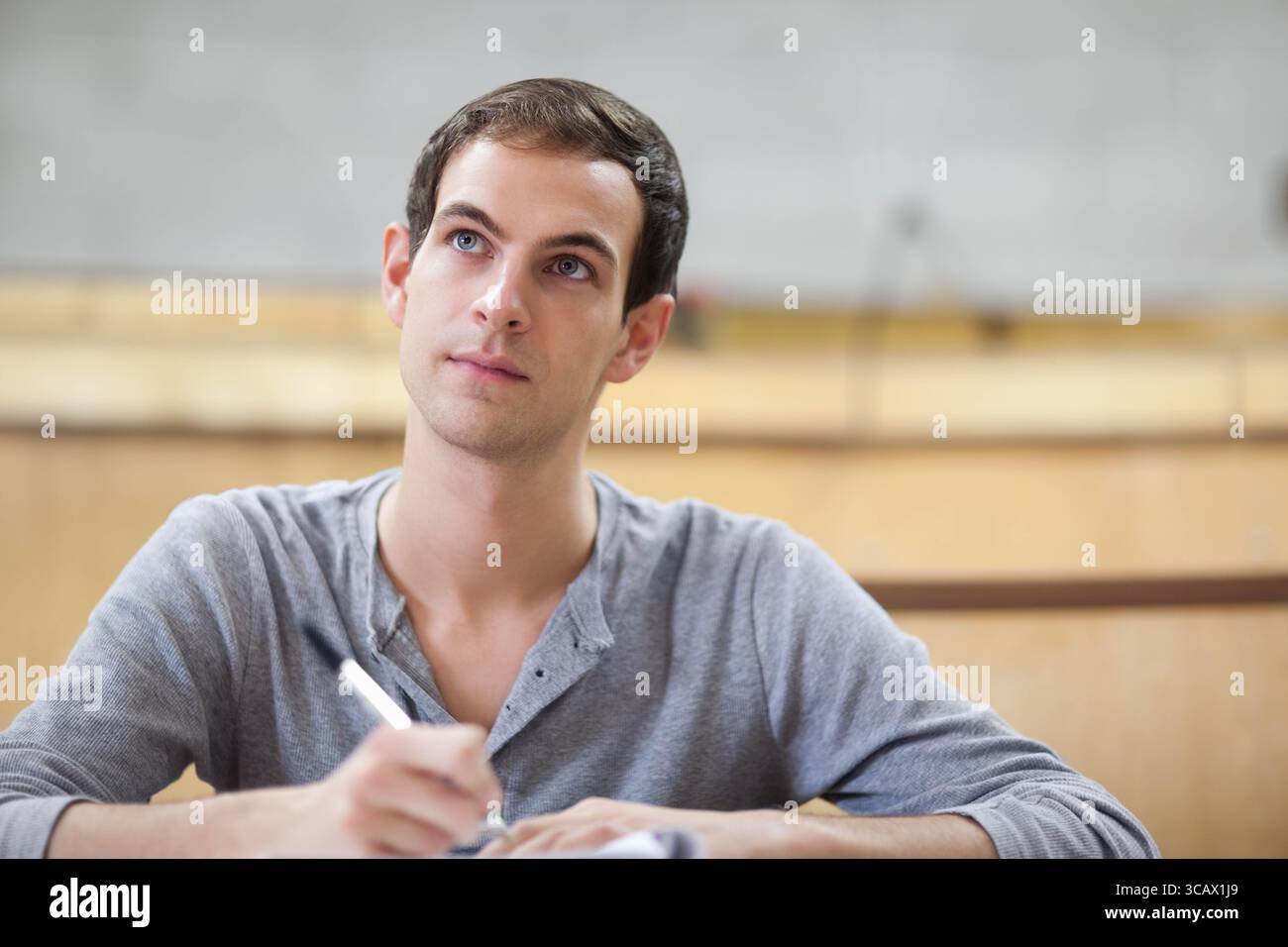 Männlicher Student sitzt am Holztisch im mehrstufigen Hörsaal und schreibt Notizen mit Stift und Notizbuch Stockfoto