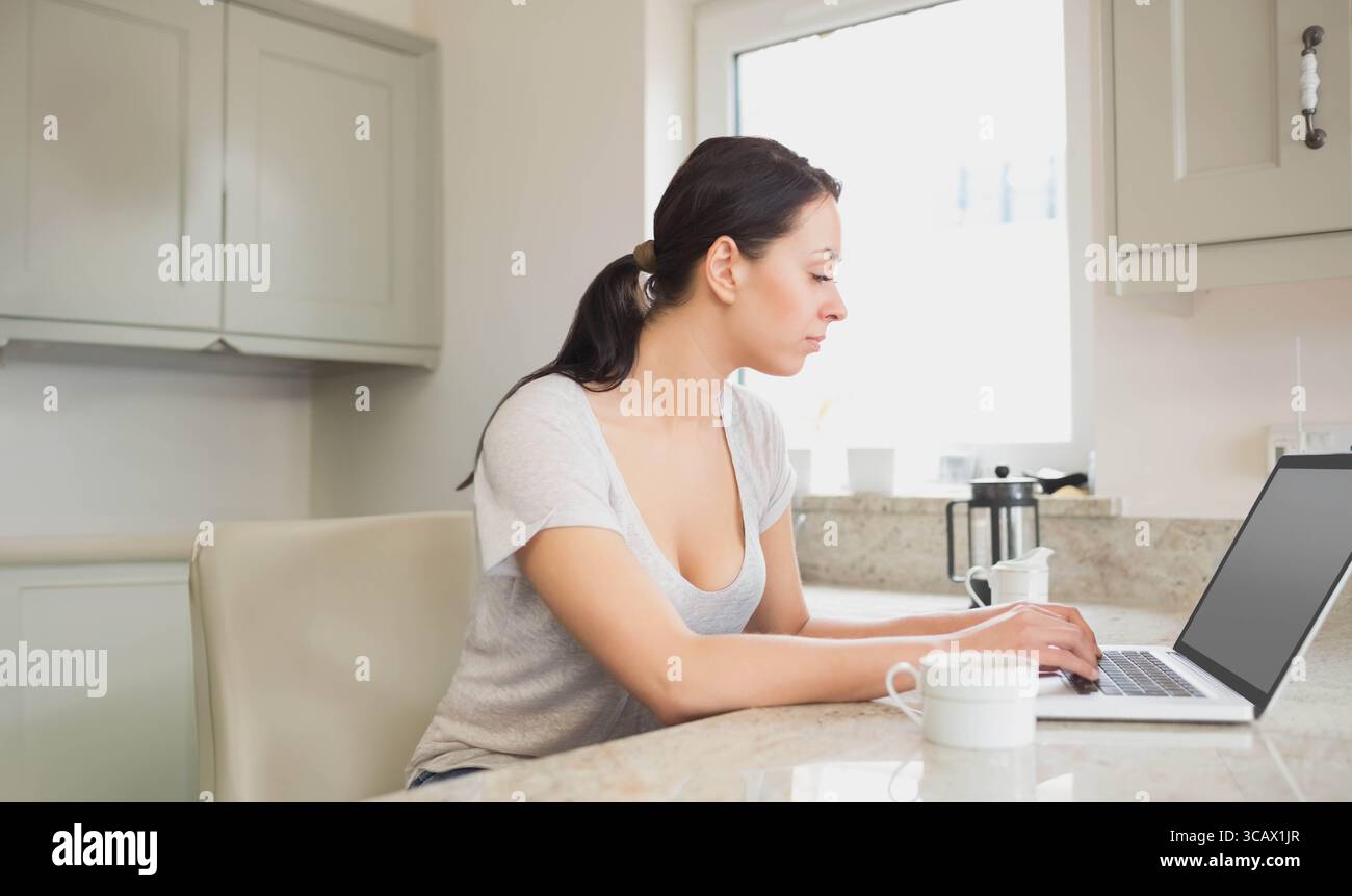 Frau, die auf einem Laptop auf der Marmor-Kücheninsel tippt, mit Kaffeetasse und Presse, Kopierraum Stockfoto