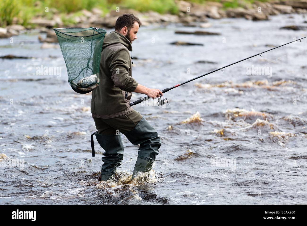 Ein Fischer überquert den Fluss mit einer Angelrute in der Hand und einem Netz, das auf dem Rücken hängt und einen Fang Fisch im Netz hält Stockfoto