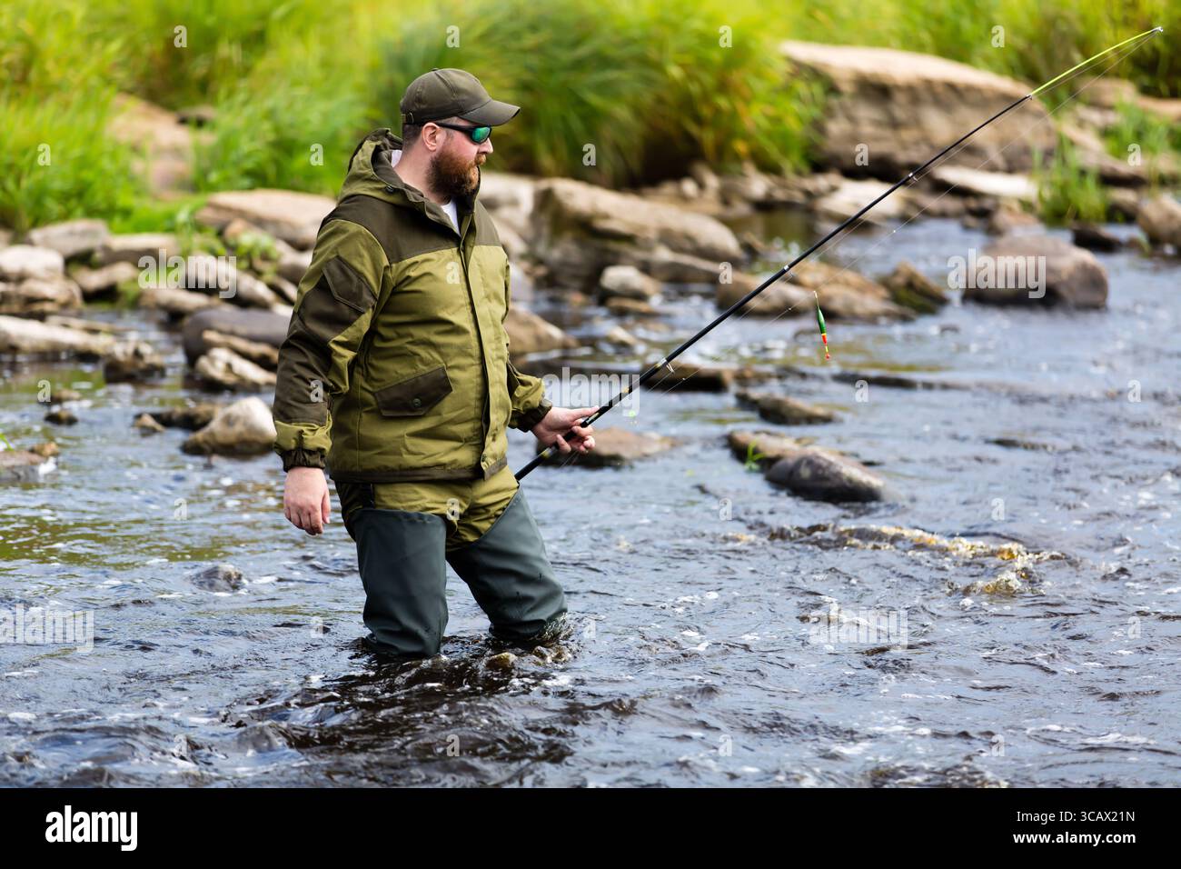 Ein harter Mann mit Bart steht im Wasser, hüfttief in Gummistiefeln und konzentriert sich auf das Fangen von Fischen in dieser Outdoor-Szene Stockfoto