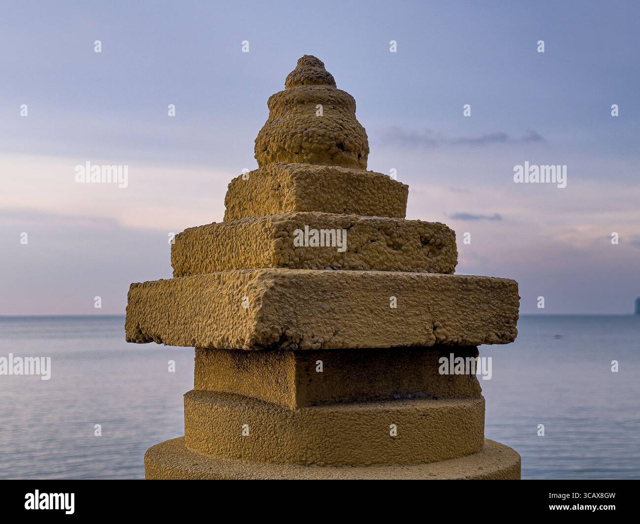 Eine verwitterte, gestufte Steinsäule mit einer abgerundeten Spitze steht vor einem ruhigen Meer und einem weichen Dämmerhimmel und zeigt ihre strukturierte Oberfläche im schwindenden Licht. Stockfoto