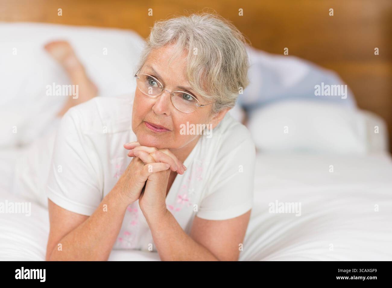 Eine ältere Frau, die auf einem Bett mit weißem Bettzeug auf Ellbogen ruht und mit Brille nach rechts blickt Stockfoto