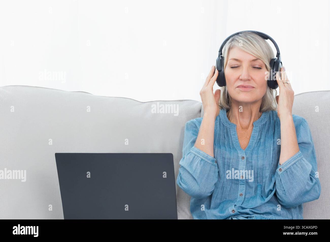 Ältere Frau, die zu Hause auf dem Sofa sitzt und Denimhemd und Kopfhörer mit Laptop trägt, Kopierraum Stockfoto