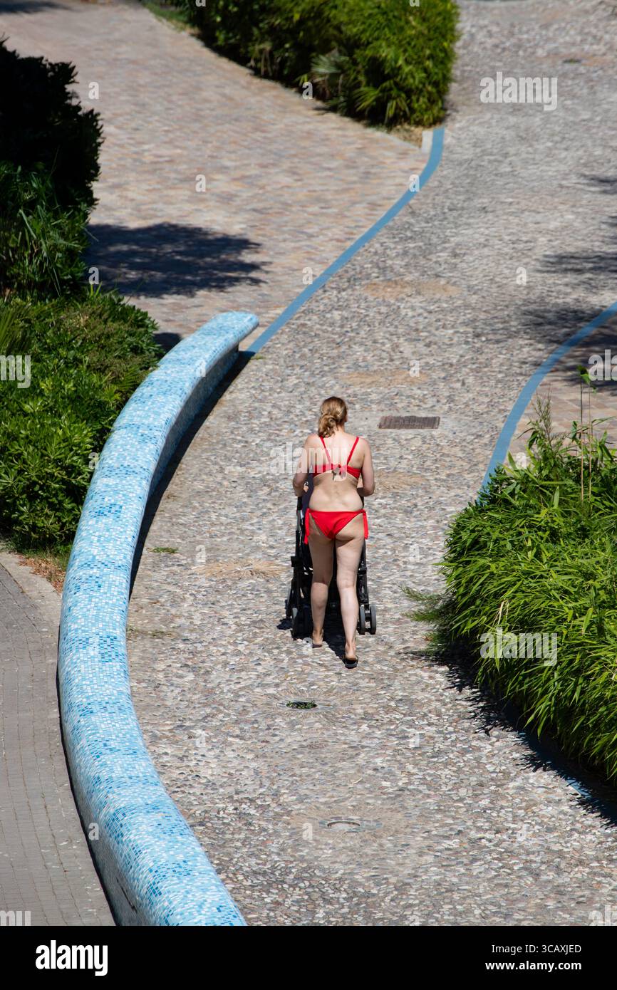 Frau schiebt Kinderwagen auf gepflasterten Weg und genießt sonnige Outdoor Walk grüne Pflanzen Sommertag. Stockfoto