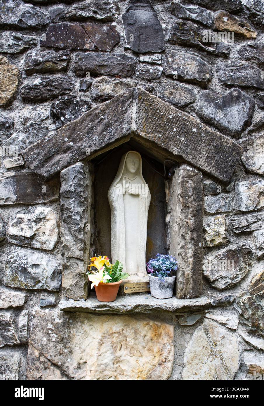 Steinstatue der Jungfrau Maria in einer rustikalen Steinmauer Nische mit bunten Blumen ein spirituelles Wahrzeichen Stockfoto