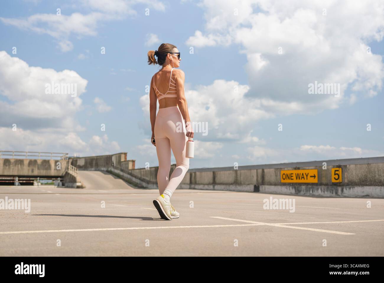 Sportliche Frau, die über einen Parkplatz läuft, Ohrstöpsel trägt und eine Trinkflasche trägt Stockfoto