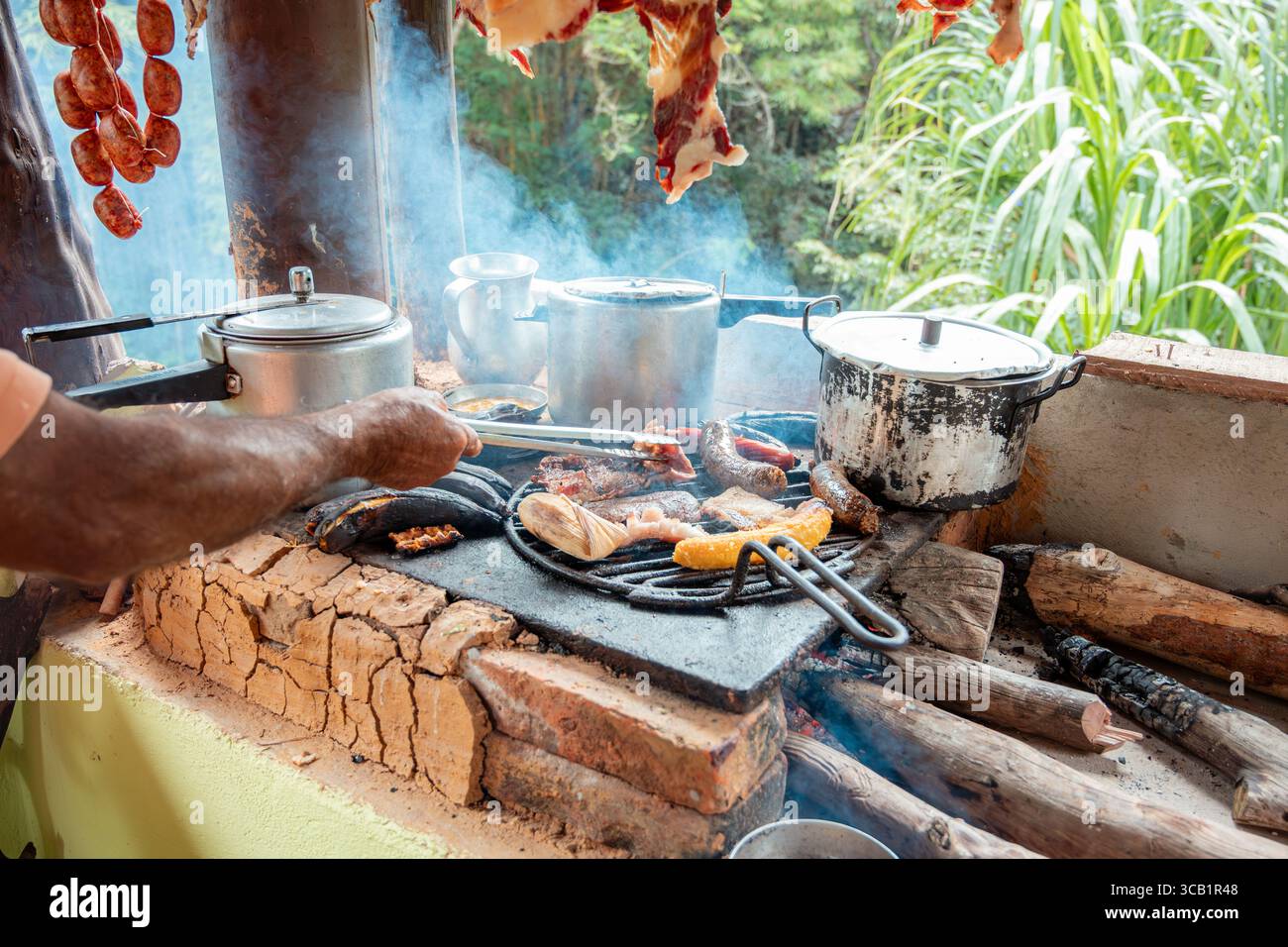 Detaillierte Nahaufnahme der Hand eines Mannes, die Fleisch auf einem rustikalen Holzofen grillt. Das Feuer, der Rauch und die Textur des Fleisches heben die Authentizität hervor Stockfoto