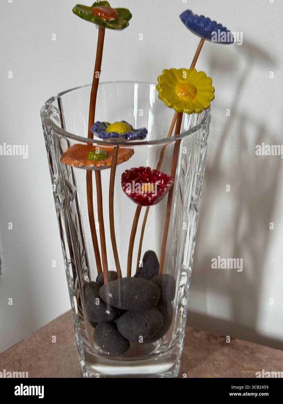 Glasvase gefüllt mit glatten schwarzen Steinen und bunten dekorativen Blumenpflücken an langen, dünnen Stielen. - Smartphone-aufgenommenes Stockfoto