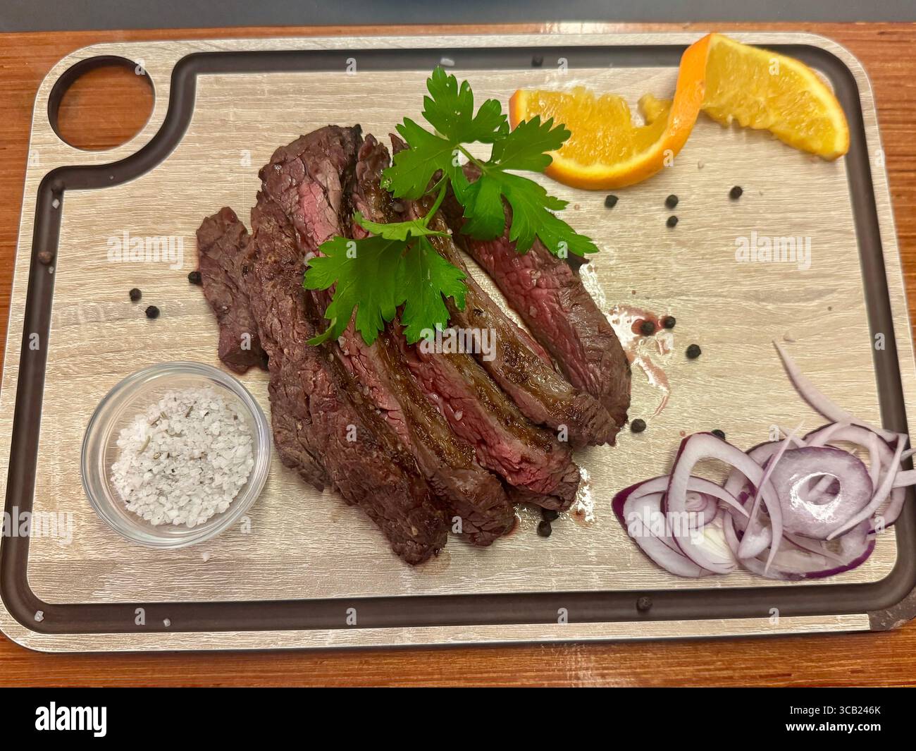 Geschnittenes gegrilltes Steak, serviert mit Meersalz, roten Zwiebeln, Orangenscheiben und Kräutern auf einem Holzbrett, präsentiert als rustikale Mahlzeit. - Smartphone-aufgenommenes Stockfoto