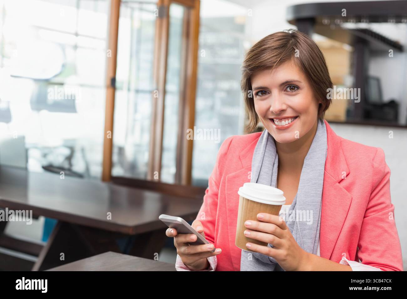Frau mit Korallenblazer, die Smartphone und To-go-Kaffeetasse am Coffee Shop-Tisch hält, Kopierraum Stockfoto